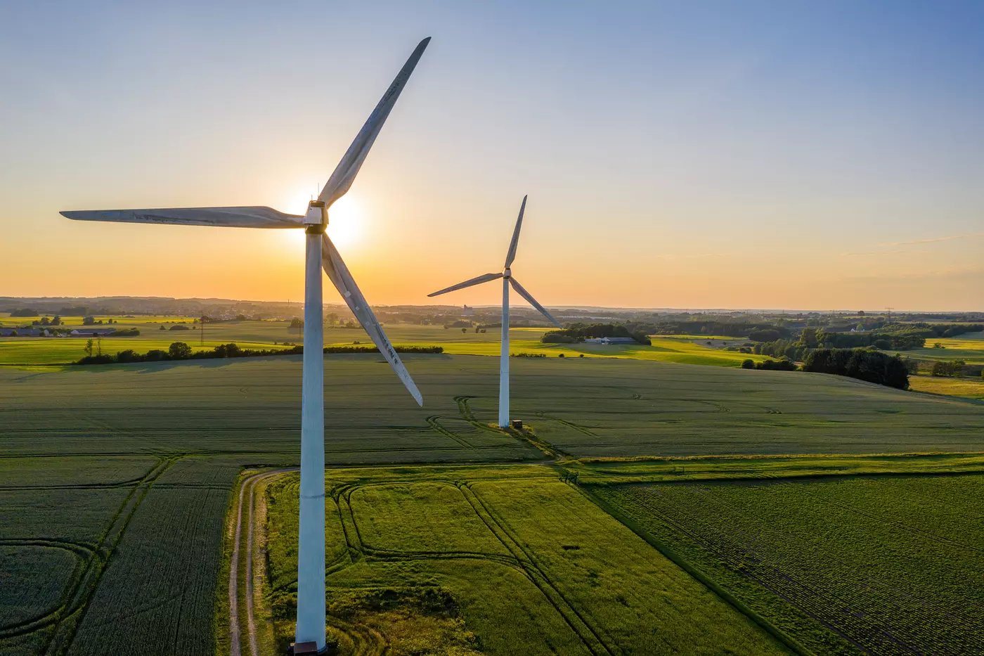 Two large wind turbines rise over flat farmland with the sun behind them low on the horizon. 