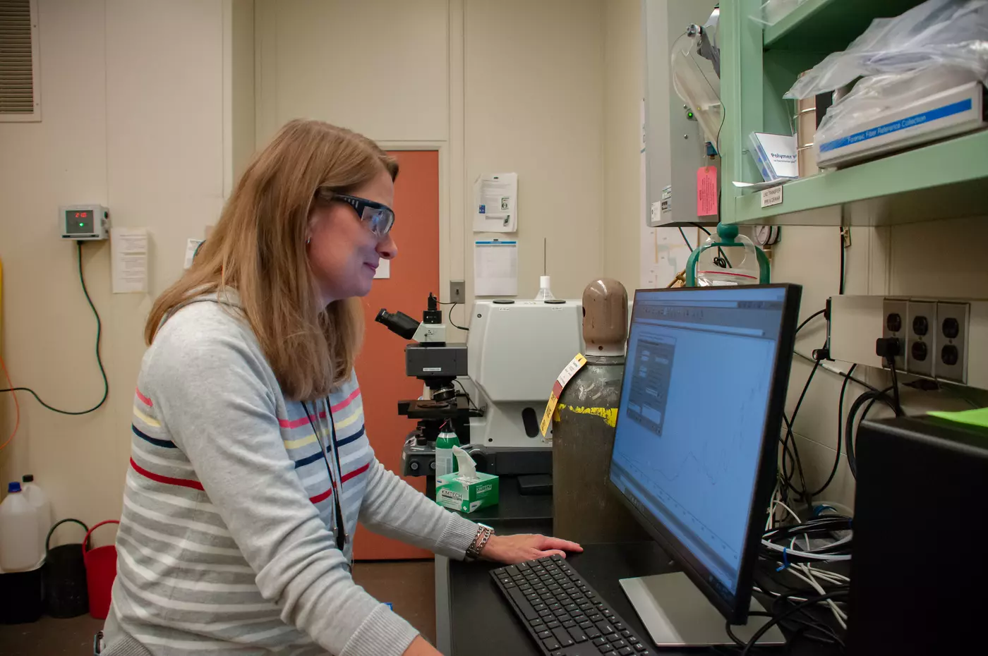 A scientist wearing safety glasses stands in front of a monitor in her lab.