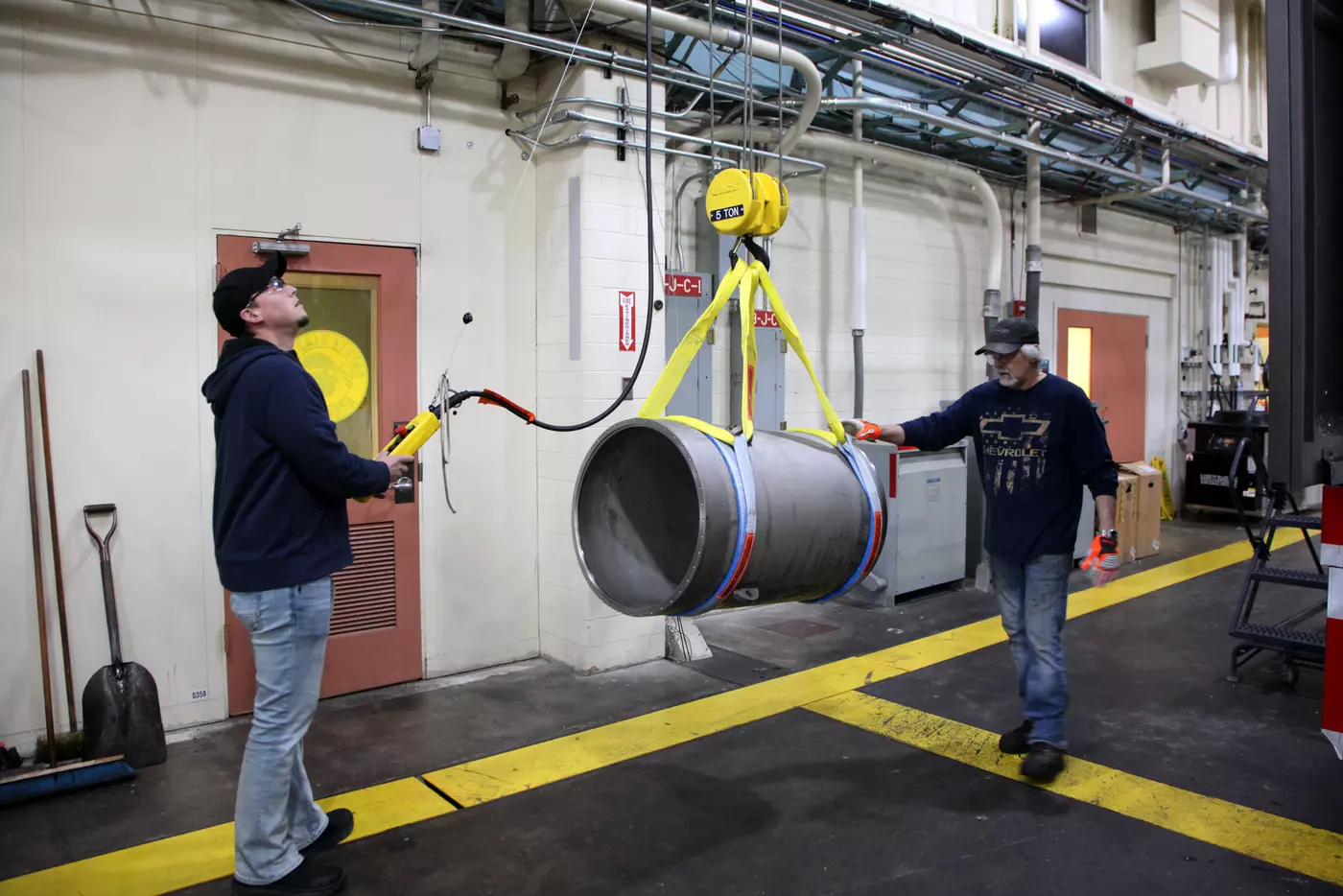 Two men use an overhead pulley apparatus to move a thick metal cylinder inside a large workshop. 