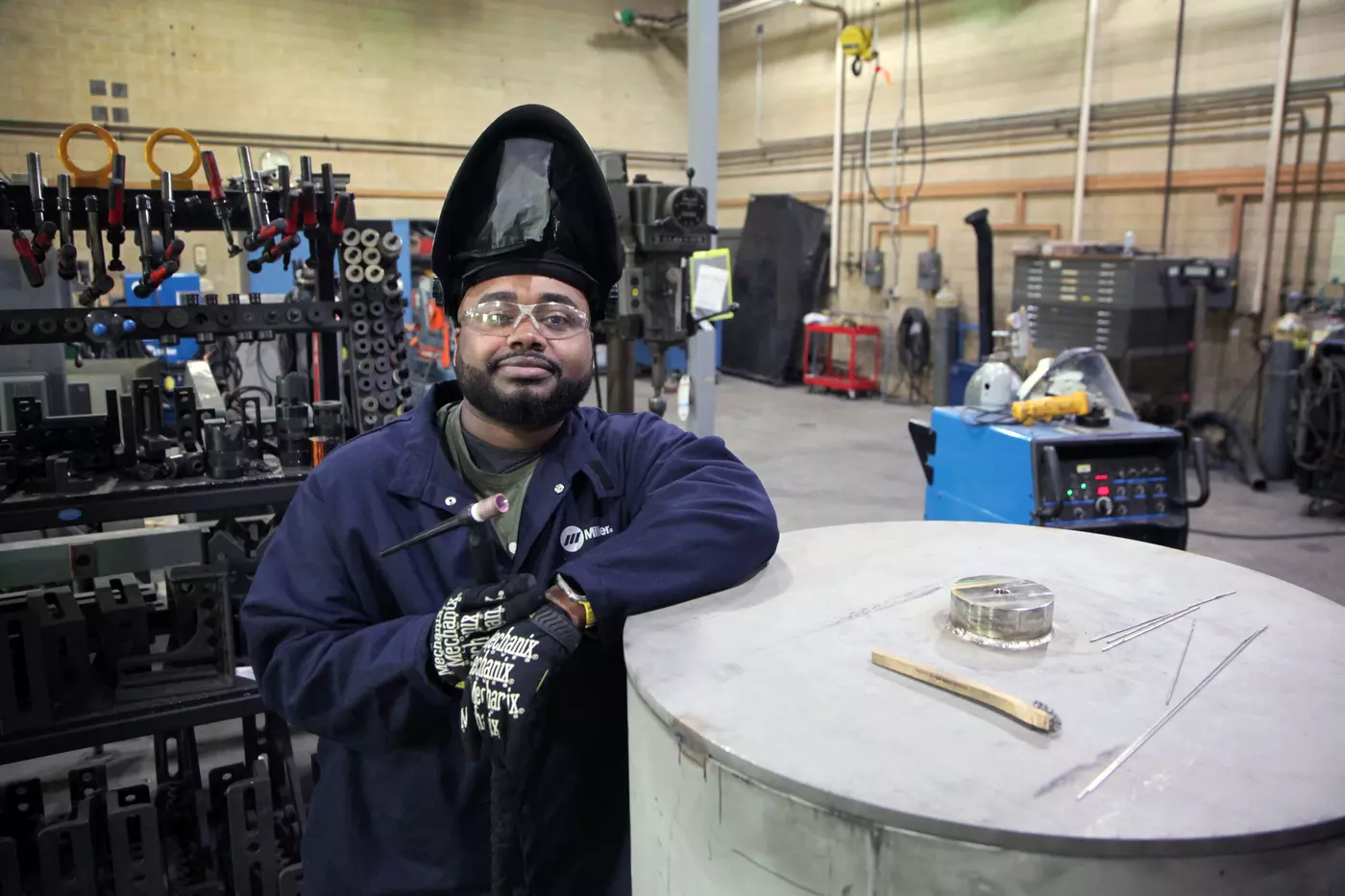A man wearing an uplifted welding mask and safety glasses poses leaning on a large metal cylinder in a workshop. 