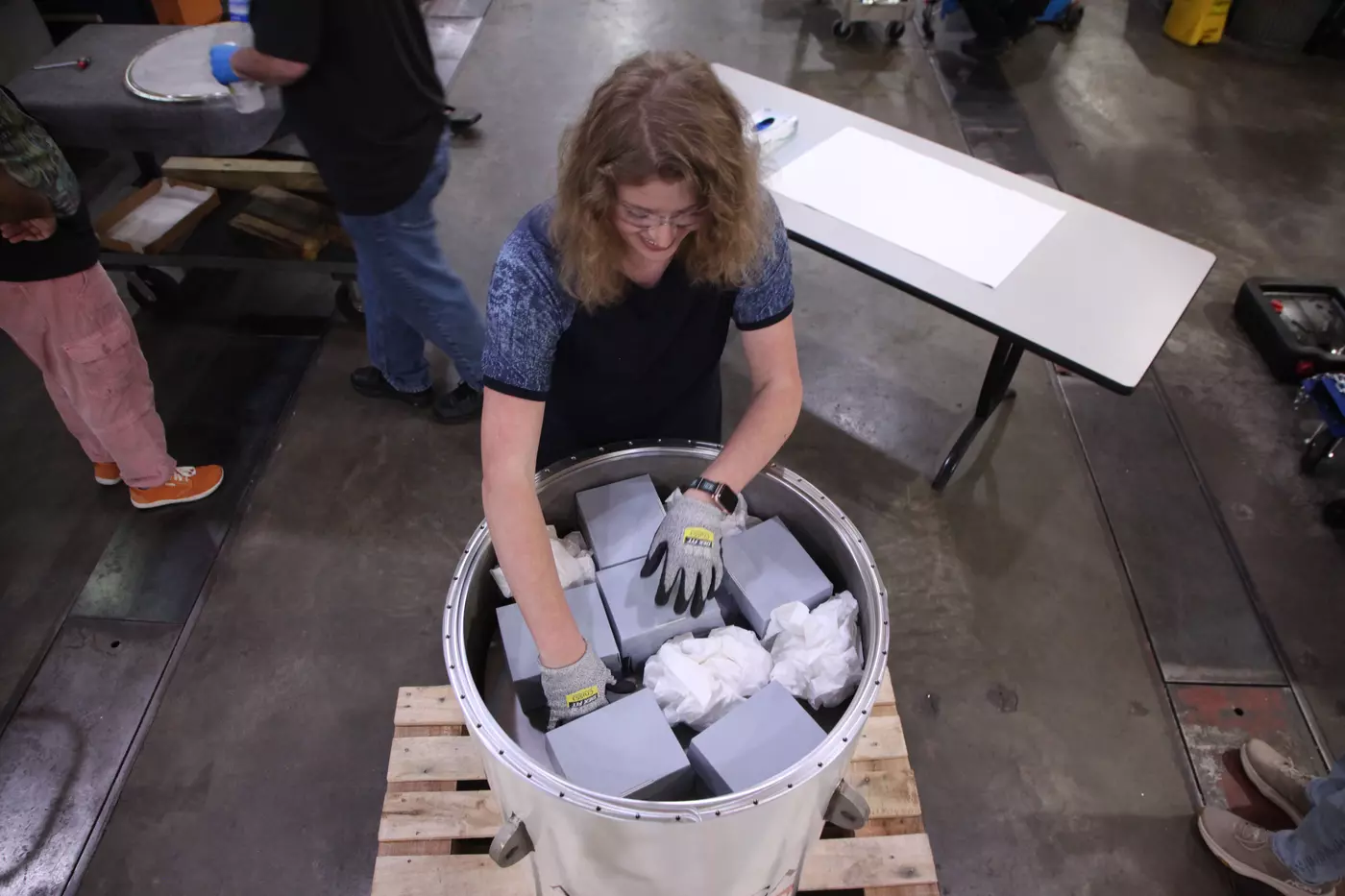 An overhead view shows a woman reaching into a cylindrical container that holds several gray boxes along with what looks like crumpled paper. 