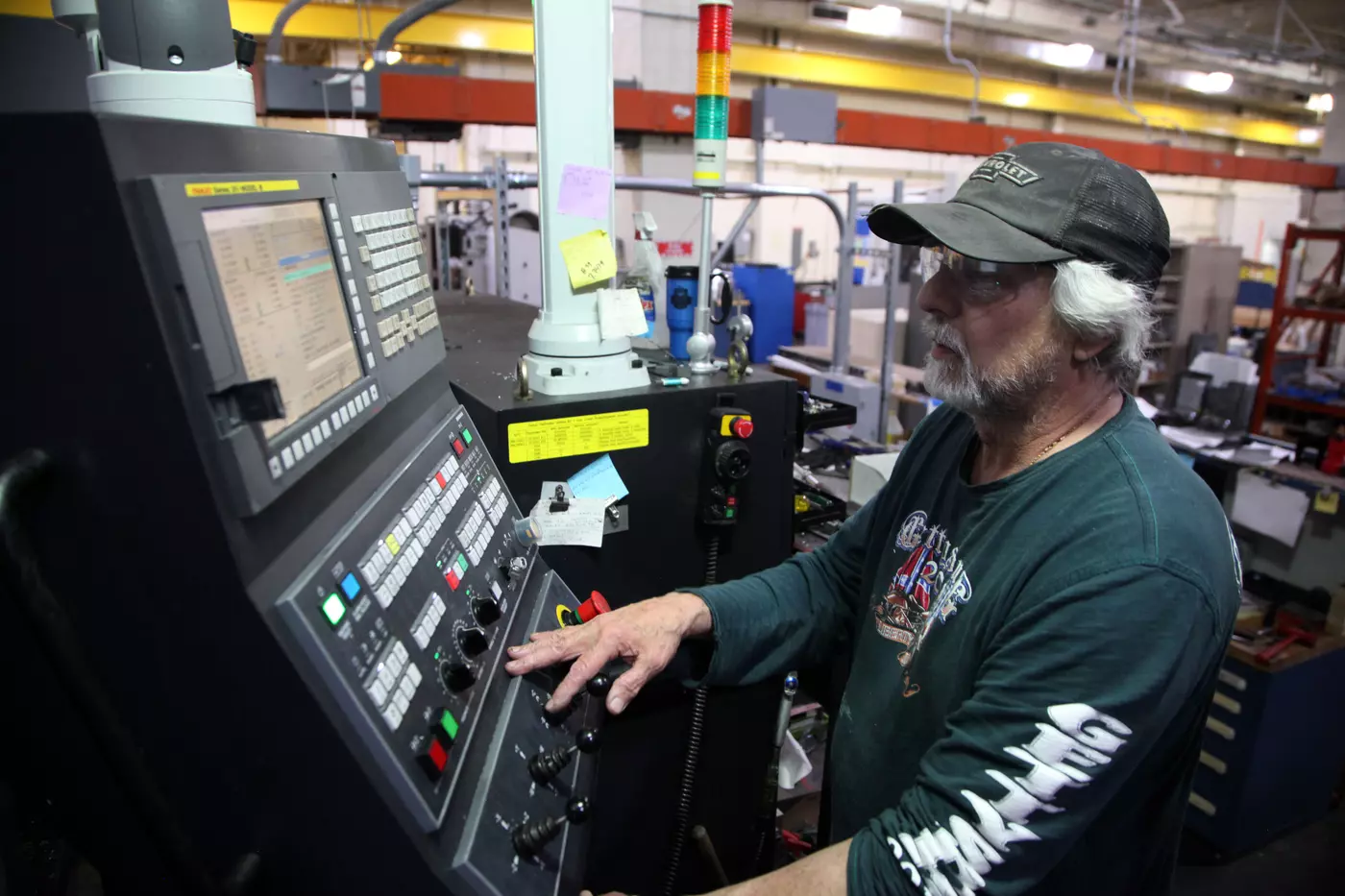 A man in a baseball cap stands at a control panel full of buttons inside a machine shop.