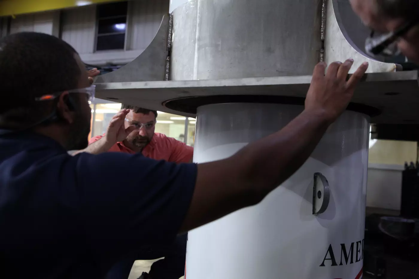 Two men reach out to hold a large metal cylinder being lowered onto a smaller one. 