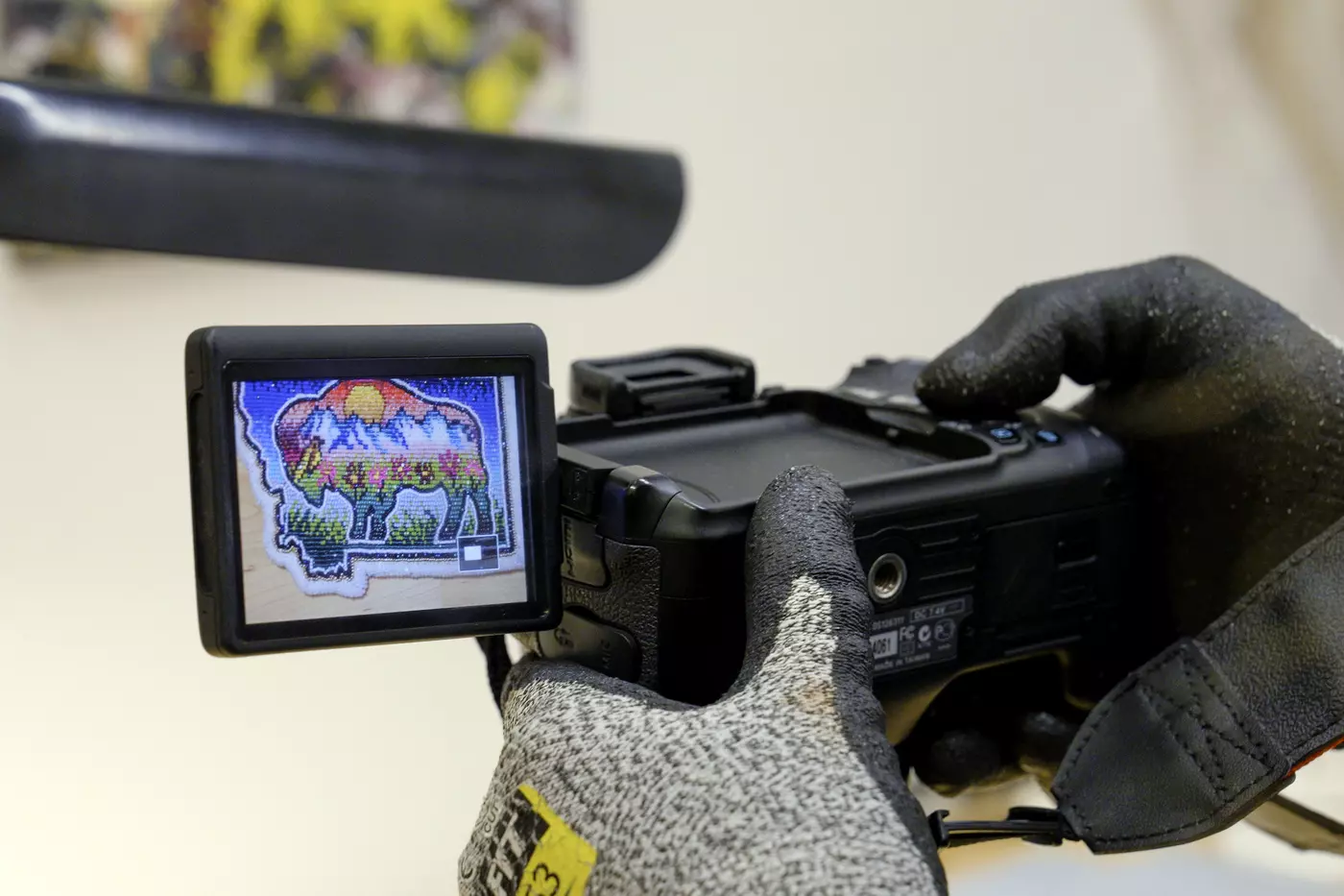 Gloved hands hold a camera; an attached screen shows a colorful beaded patch depicting a buffalo. 