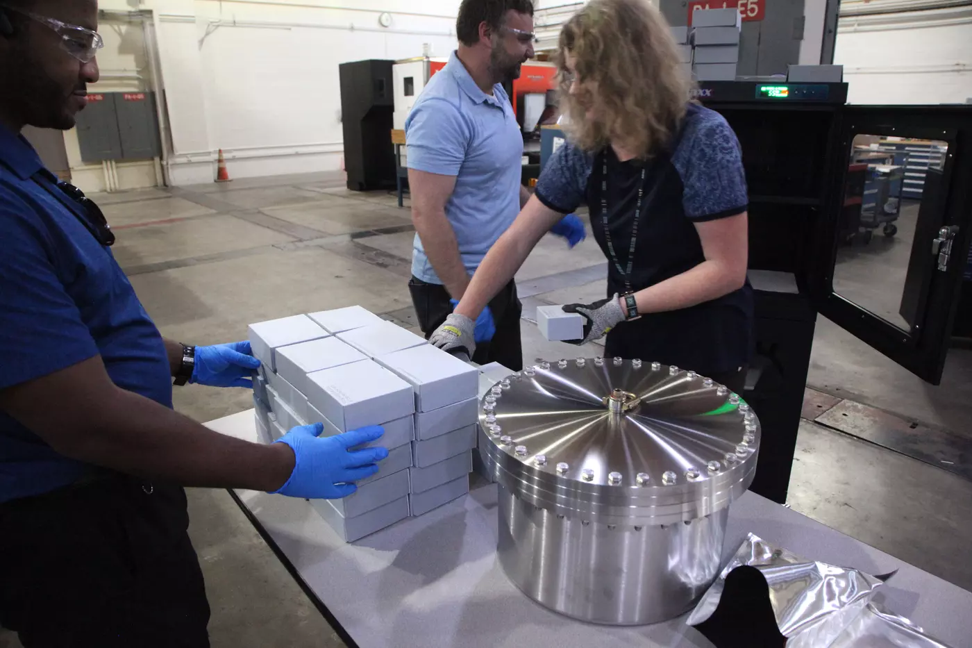 A woman wearing gloves stacks identical small boxes on a table in a machine shop, with other workers around her to help.