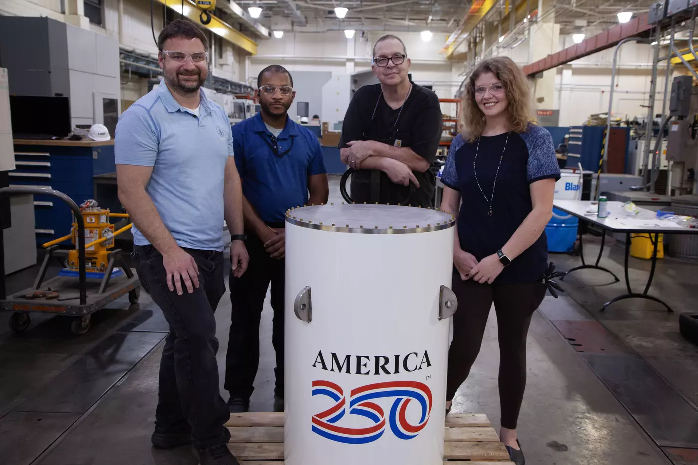 Four workers in a machine shop pose smiling around the upright white cylinder of the America250 time capsule. 