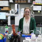 Briana Capistran wears safety glasses as she poses smiling in the lab with computers and other equipment in the background. 
