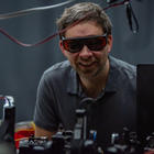 Krister Shalm, wearing safety glasses, smiles over a lab table covered with electronics equipment.