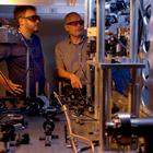 Two researchers in safety glasses stand behind a table holding various electronic and laser devices. A metal structure in the foreground is part of the atomic clock. 