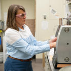 A researcher wearing safety glasses stands in the lab, reaching into a metal device on the lab table. 