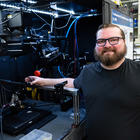 A bearded man poses smiling next to an open cabinet of equipment in a lab. 