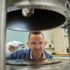 Andrew Iams wears safety glasses as his face is framed in an opening between two horizontal pieces of equipment in the lab. 
