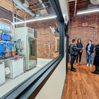 Four people stand talking in a converted factory space near a large interior window through which a complex, high-tech manufacturing line is visible. 