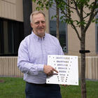 Jack Glover poses outside on the NIST campus, holding a flat square object with holes of varying sizes. 