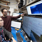 A woman with glasses and a long ponytail is seated at a computer with four display screens. A man with glasses stands beside her and points at the uppermost display screen. In the middle of that screen is a small blue glowing dot. The lower screens display data from the experiment.