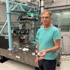 Jim Cline stands in front of a large scientific device in a lab, holding a small white disk.