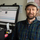 Gary Howarth poses for a photo in his office, wearing a NIST baseball cap, with a ruggedized laptop on the counter next to him and a map on the wall behind him.