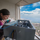 A researcher peers into a box of scientific equipment near an opening in the wall showing the view down the mountain. 