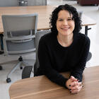 Kelly Sauerwein poses sitting at a table in the NIST library, leaning forward and smiling. 