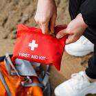 A person holds a red pouch labeled First Aid Kit as they stand over a travel bag on sandy ground.