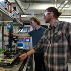 Two male researchers in casual clothes and protective eyewear stand in the lab, pointing at a screen hooked up to a laser science instrument. 