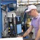 A man wearing safety glasses and a baseball cap leans over a lab table holding computer monitors that show spreadsheets of data.