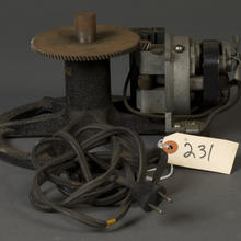 geared mechanism with an electric plug. Attached to the plug's wire is a tag that reads "231." The device is geared to another smaller gear on a device with what looks like a flywheel coming from the front of it. 