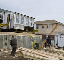 Men working at construction site with wood planks on ground and gray house dangling in air by crane