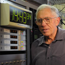 Man with gray hair standing beside computer with digital clock display