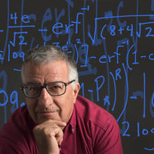 Former NIST researcher Tassos Nakassis sitting in front of a black background. The background is covered in blue equations.