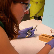 Woman with safety glasses and purple gloves working in lab. In her left hand she has a device with a tube attached to it. The tube is running into a small vial. In her right hand she holds a coiled tube leading from the vial.