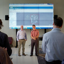 Two men stand in front of an audience, demonstrating WIPP, which is projected via computer onto several large screens in a library.