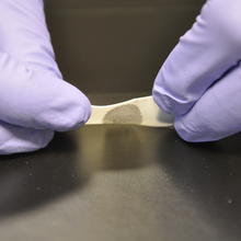 A close up of two purple gloved hands removing a piece of tape from a shiny surface. A fingerprint can be seen on the tape.