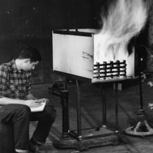 Black and white photo of a young man hunched over sitting on a chair next to a large white box that is burning a wood fire for an experiment.