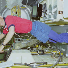 Color photograph of man wearing red shirt and bluejeans floating inside space shuttle. The space shuttle silver walls are covered in yellow pipes and red buttons from floor to ceiling.