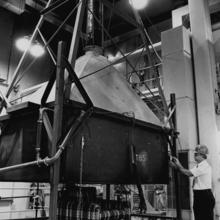 Black and white photo of a man standing in front of large piece of metal equipment shaped like a funnel. It reaches from the floor to the ceiling.