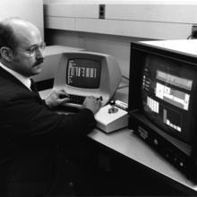 Black and white photo of man working on two computer screens in 1989. The screen to the left is small and long and the screen to the right is large and boxy, both show correlating data. 