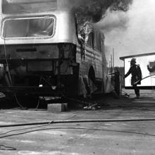 Black and white photo of bus on fire surrounded by a firemen carrying a water hose in 1975. 