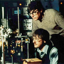 Bill Phillips standing in his lab with postdoc John Prodan seated.