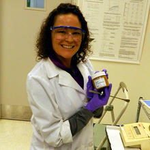 NIST chemist Carolyn Burdette in a white lab coat and wearing goggles holding up a jar of  SRM 2387, Peanut Butter