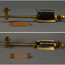 A brass rod with a back box mounted to a brass plate on one end and a thin brass plate stuck perpendicularly to the rod on the other. There is a lever on the black box and an unattached rectangular coil of copper wire resting next to the device. 