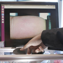 A finger presses on a fingerprint scanner. In the background, the fingerprint is shown on a computer monitor.