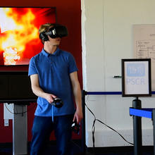 Person wearing a VR headset and holding controllers. Behind him a screen shows what the researcher is seeing within the headset--a virtual office building that is on fire.