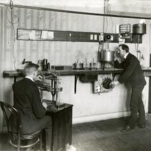 Man sits at a desk on the left while a man on the right stands looking into a device