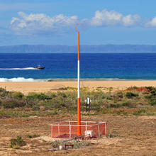 An orange and white antenna a few meters away from a beach. A boat speeds past in the distance. Beyond that the island of Niihau is visible. 
