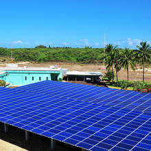view of an array of blue solar panels with a turquoise building behind them on a barren patch of soil. The beach and ocean are in the distant background. 