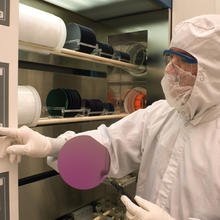 Man wearing a white protective coverall, hood, gloves and face mask, holds a round, purple, sample of a silicon wafer and presses buttons on a white instrument panel.