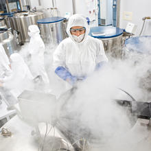 Giant metal tubs are being opened and white vapor is puffing up into the face of one researcher who is in a protective suit with hood, gloves and eye protection. In the background, several other suited researchers discuss something.