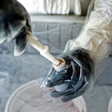 A pair of gloved hands prepares a specimen for storage using a test tube and a special, slim scoop.