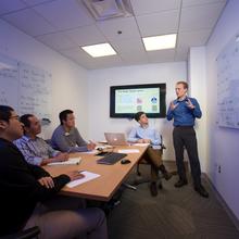 man standing in front of a white board filled with equations. Four other men sit around a table watching.